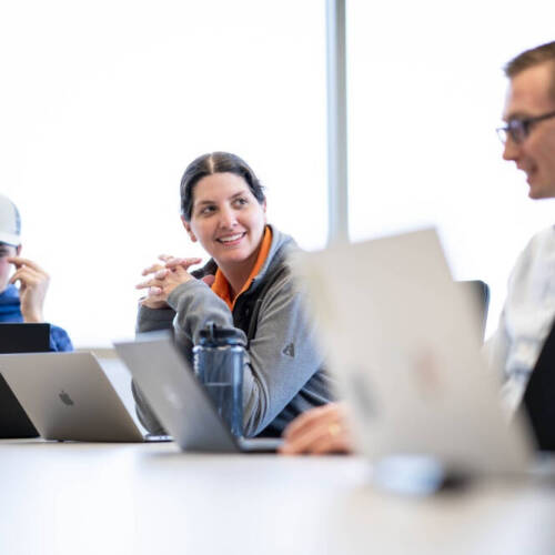 Three people are seated around a conference table, each working on laptops. One person is wearing a cap with the letters "GV" on it. The setting appears to be a modern meeting room with large windows in the background, allowing natural light in.